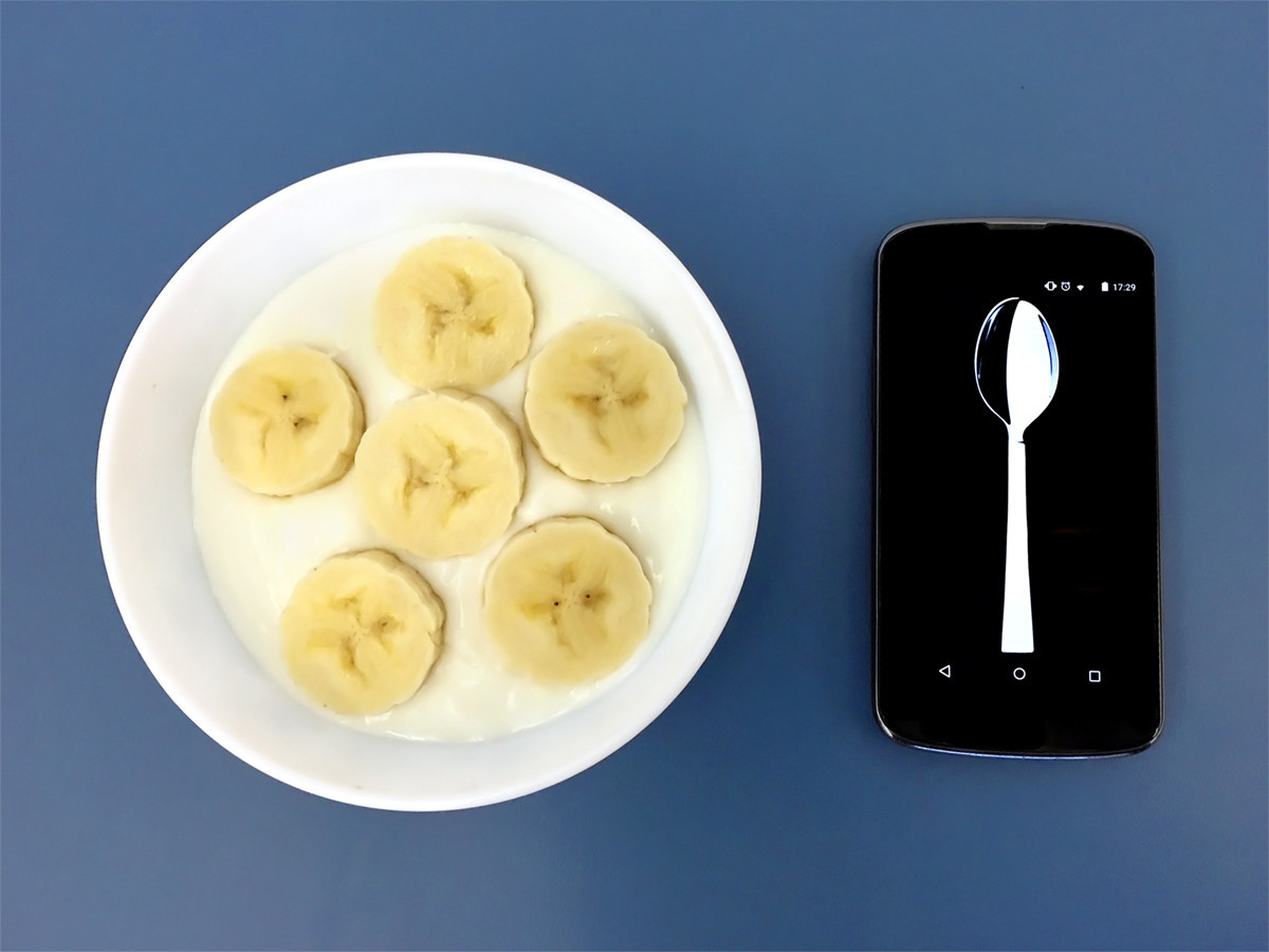 A phone displaying a spoon beside a bowl of yogurt and banana slices on a blue surface